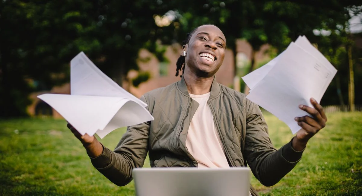 Cheerful busy black man in casual clothes and wireless earphones holding documents on sheets of paper while sitting in park w