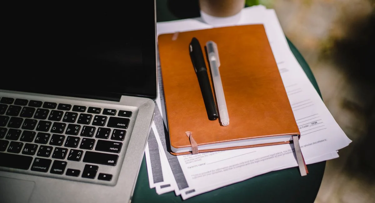 From above of leather notebook with papers and pens placed on table near laptop and cup of coffee