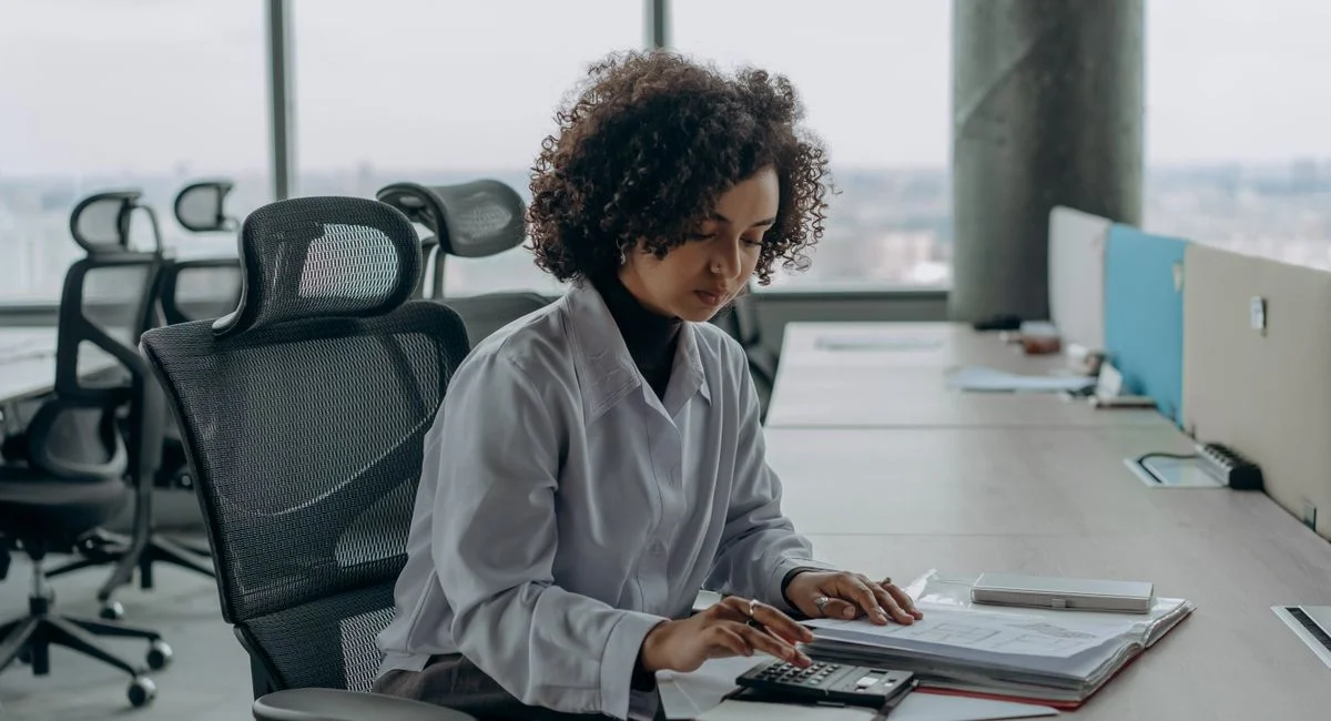 A professional woman with curly hair calculating documents in a modern office setting.