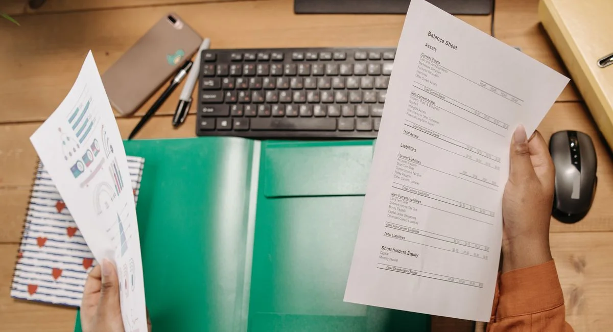 Hands holding and reviewing balance sheets over a desk with a keyboard and stationery.
