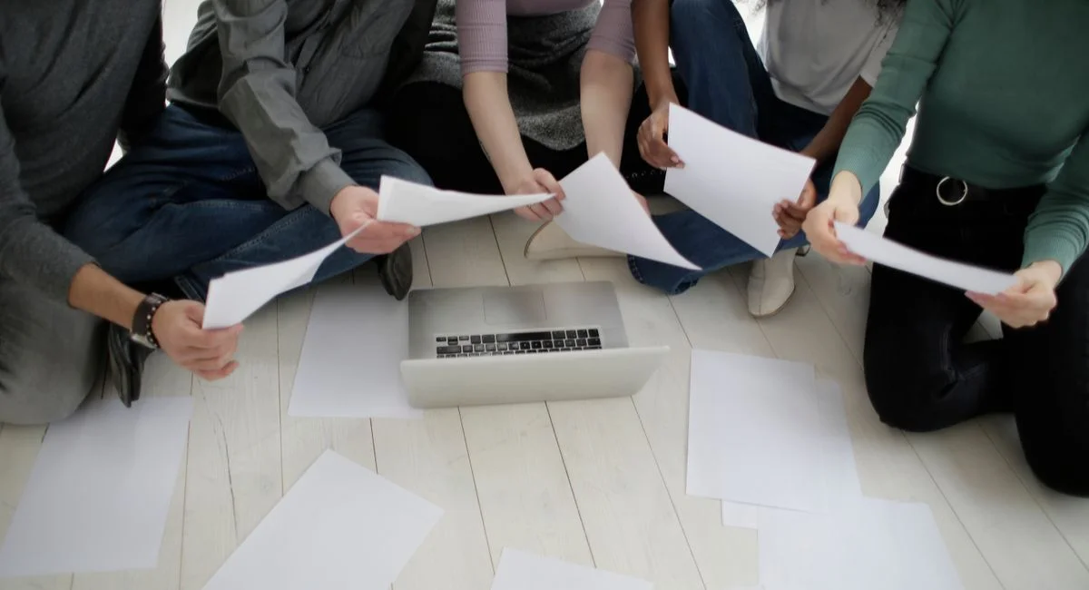 Crop faceless diverse coworkers in casual wear collecting assorted empty white papers sitting on floor in front of netbook in