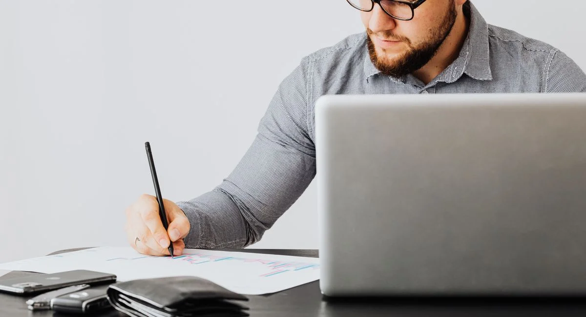 A focused man analyzing financial papers with a laptop in an office setting.