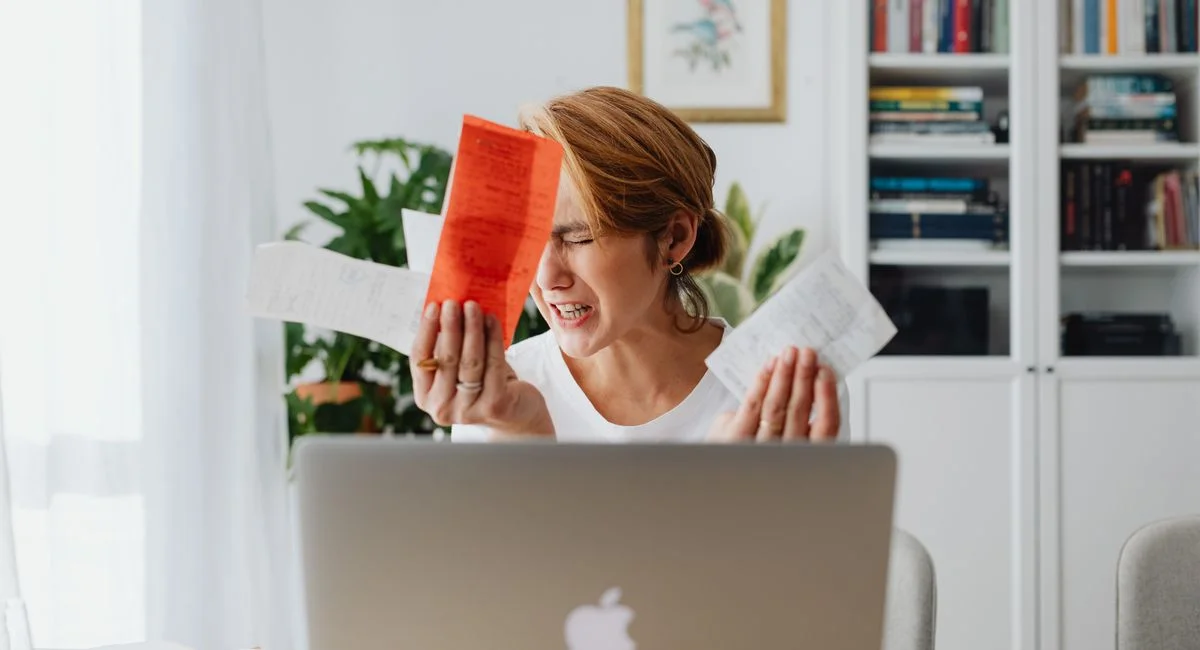 Frustrated woman managing finances, surrounded by bills and a laptop, looking distressed.